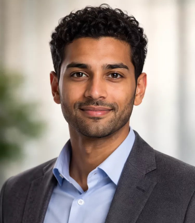 A man wearing a gray suit jacket and light blue dress shirt is pictured in a professional setting. The background is softly blurred, featuring neutral tones and hints of greenery. The overall mood is polished and business-oriented.